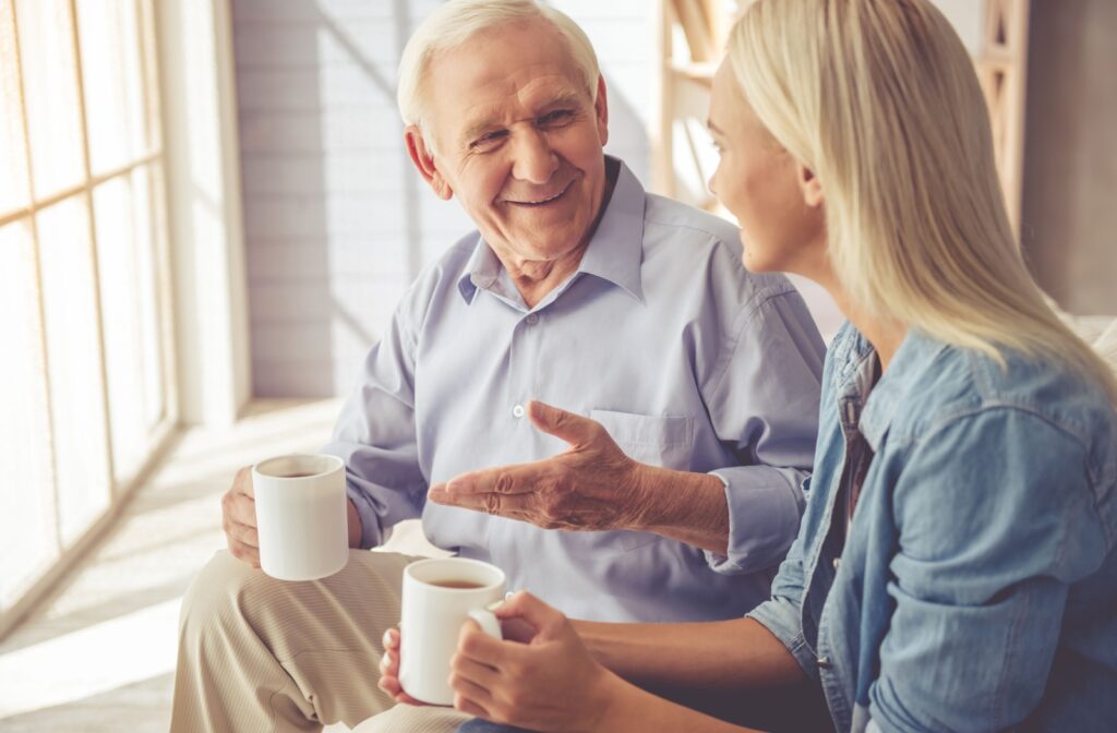 Older man and younger woman sitting and chatting while drinking out of mugs.
